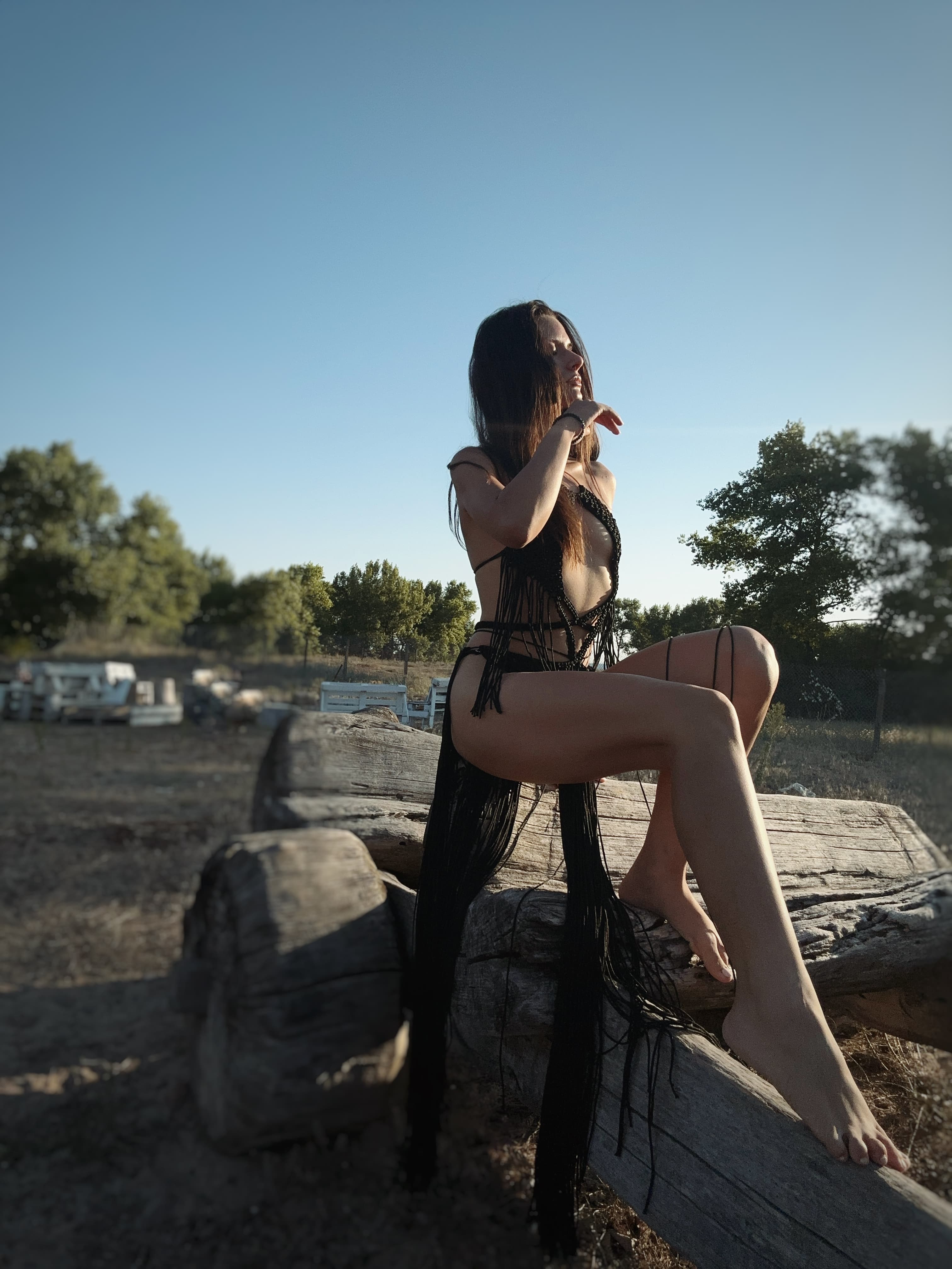 A woman wearing a black crochet maxi dress sits on a wooden log outdoors under a clear blue sky.